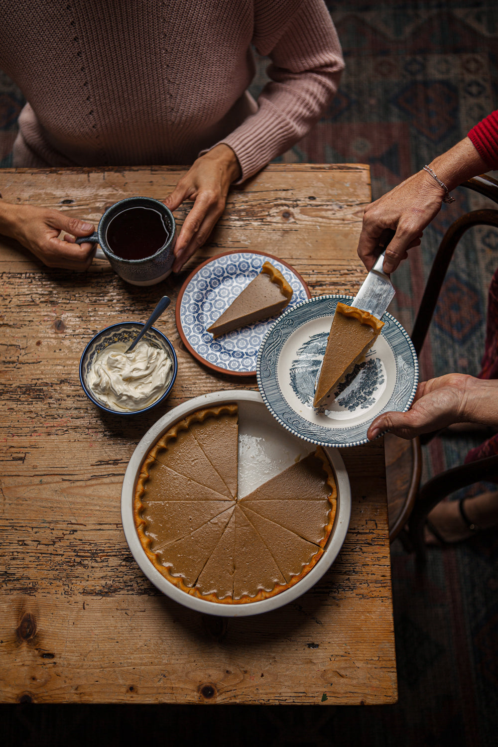 Pumpkin pie with slices on a wooden table, accompanied by a mug of coffee and a bowl of whipped cream.