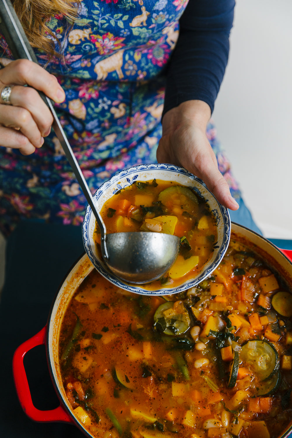 Person ladling vegetable soup from a pot with a colorful patterned apron.